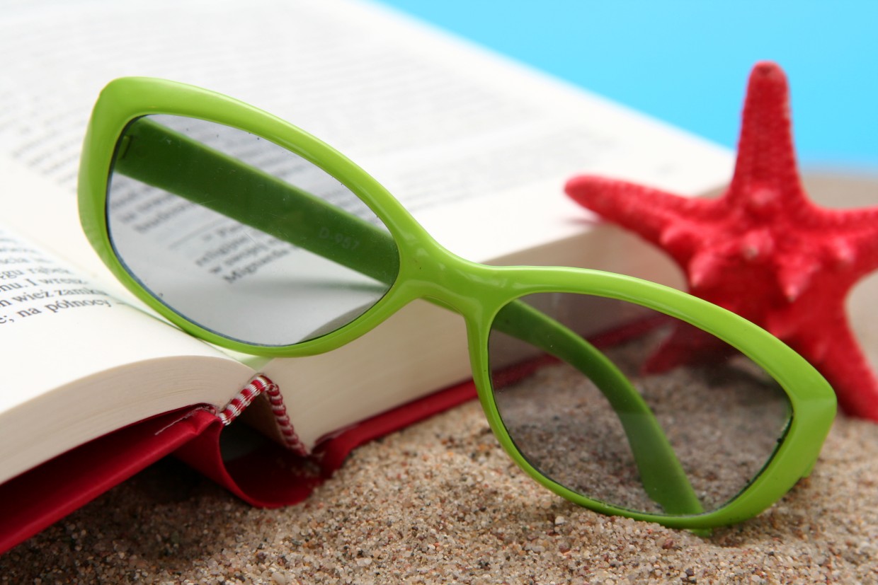 A book, starfish, and pair of sunglasses sit atop sand and an ocean blue background.