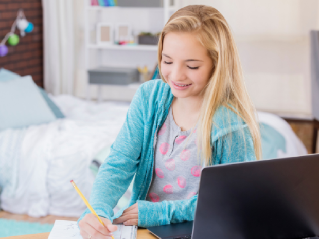 A tween girl smiles down at her notebook, a pencil in her hand a laptop open in front of her.