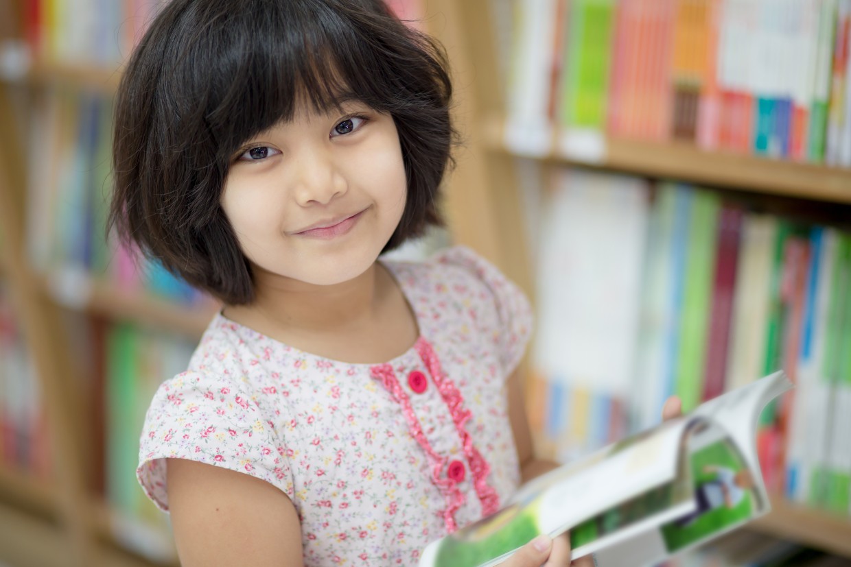 A child holds an open book and stands in front of full bookshelves.