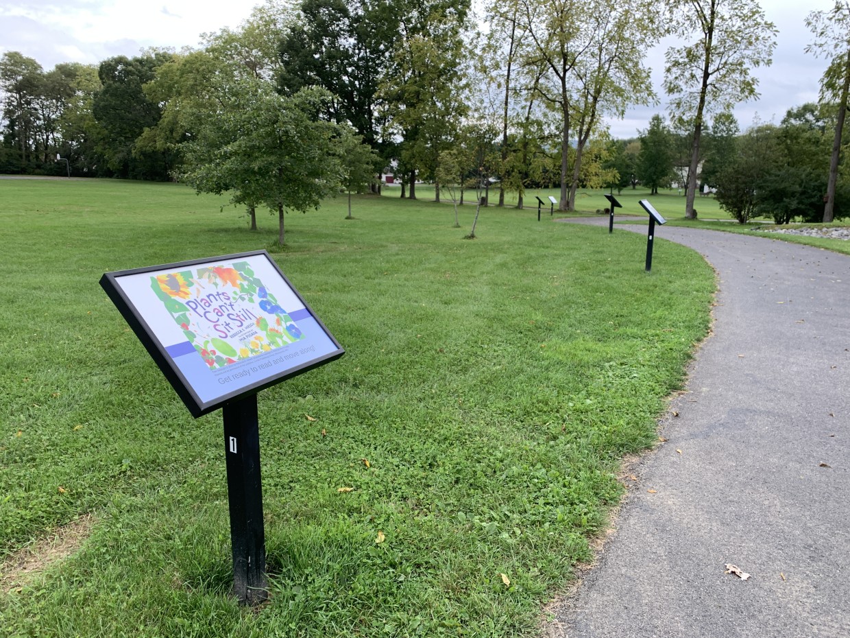 Photo of a frame on a post along a walking path in Autumnwood Park, displaying the cover of the book Plants Can't Sit Still by Rebecca Hirsch.