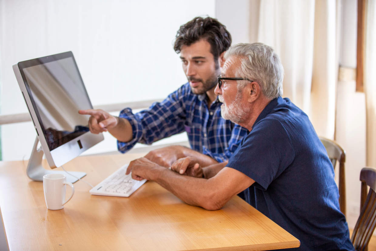 Younger man pointing to computer screen, sitting with older man typing on the same computer.