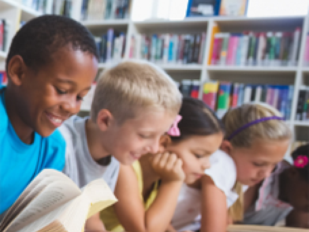 A group of children lie on the floor, open books in front of them and bookshelves behind them.