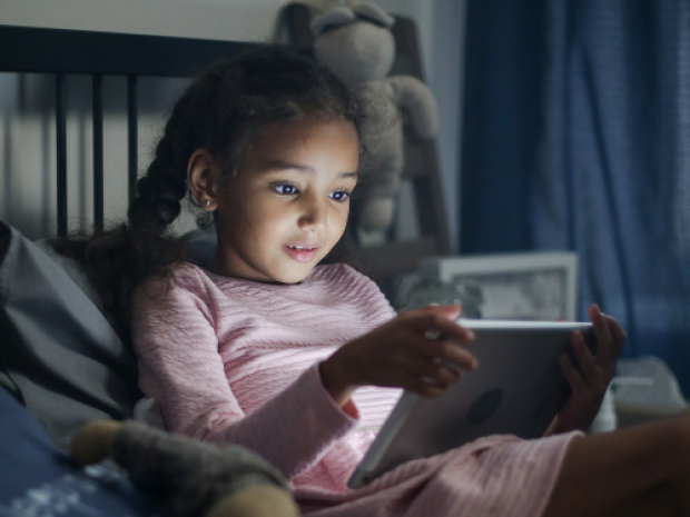 A child sits in their bed in the dark, face illuminated by the tablet they hold