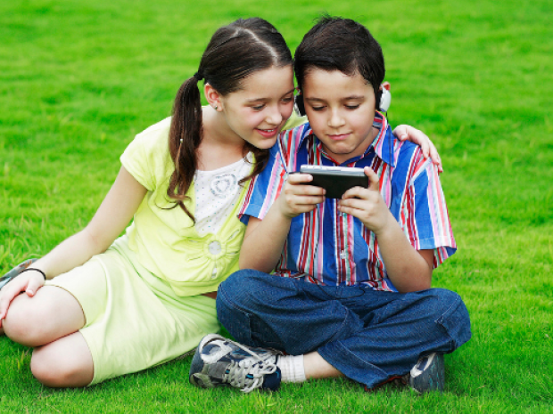Two children sit in a field of grass, leaning together and looking at a phone