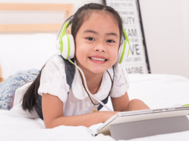 A child lays on their stomach on their bed, a tablet in front of them and headphones on their ears