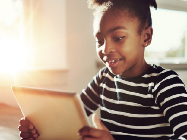 A child sits at a table holding a tablet in their hands, sunlight coming through the window behind them