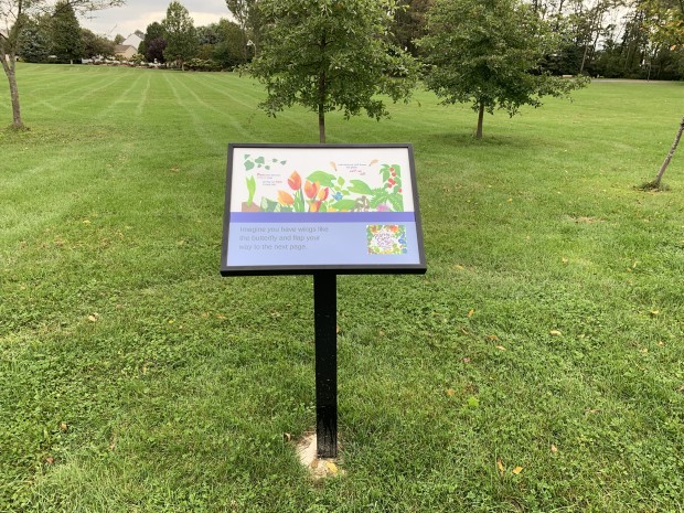A StoryWalk post - a black frame displaying two pages of a picture book - stands in the grass of a local park
