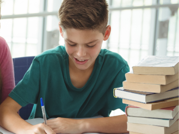 A child sits next to a stack of books, writing in an open notebook in front of them