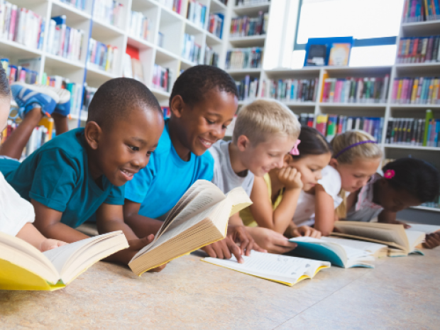 A group of children lie on the floor on their stomachs, looking at books, with full bookshelves behind them